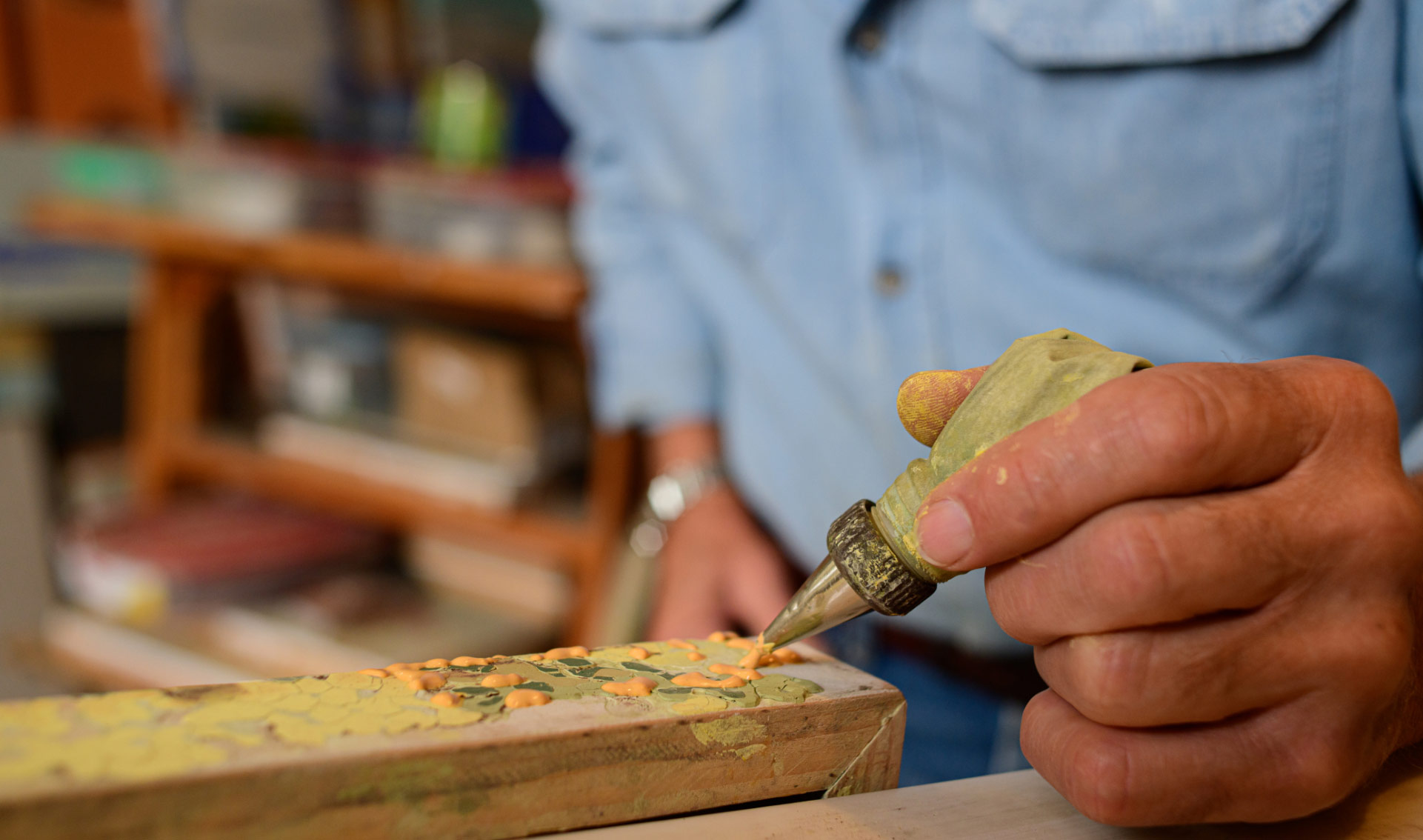 A close-up of Lynn’s hand, holding a piping bag with a metal tip on it. A thick orange substance that looks like paint is being squeezed out of the piping tip onto the edge of a canvas.