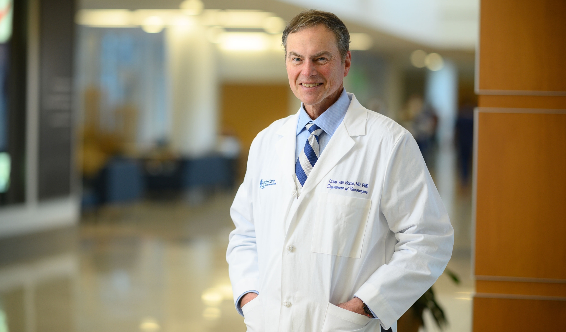 A photo of Dr. Craig van Horne posing in the hallway at UK HealthCare. He is an older white man with short gray hair, wearing a white lab coat over a blue button-up and a blue and white striped tie.