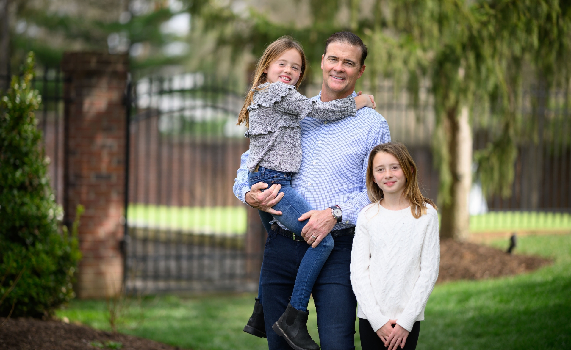 A photo of Coach Mitchell posing outside with his daughters, one of whom he is holding.