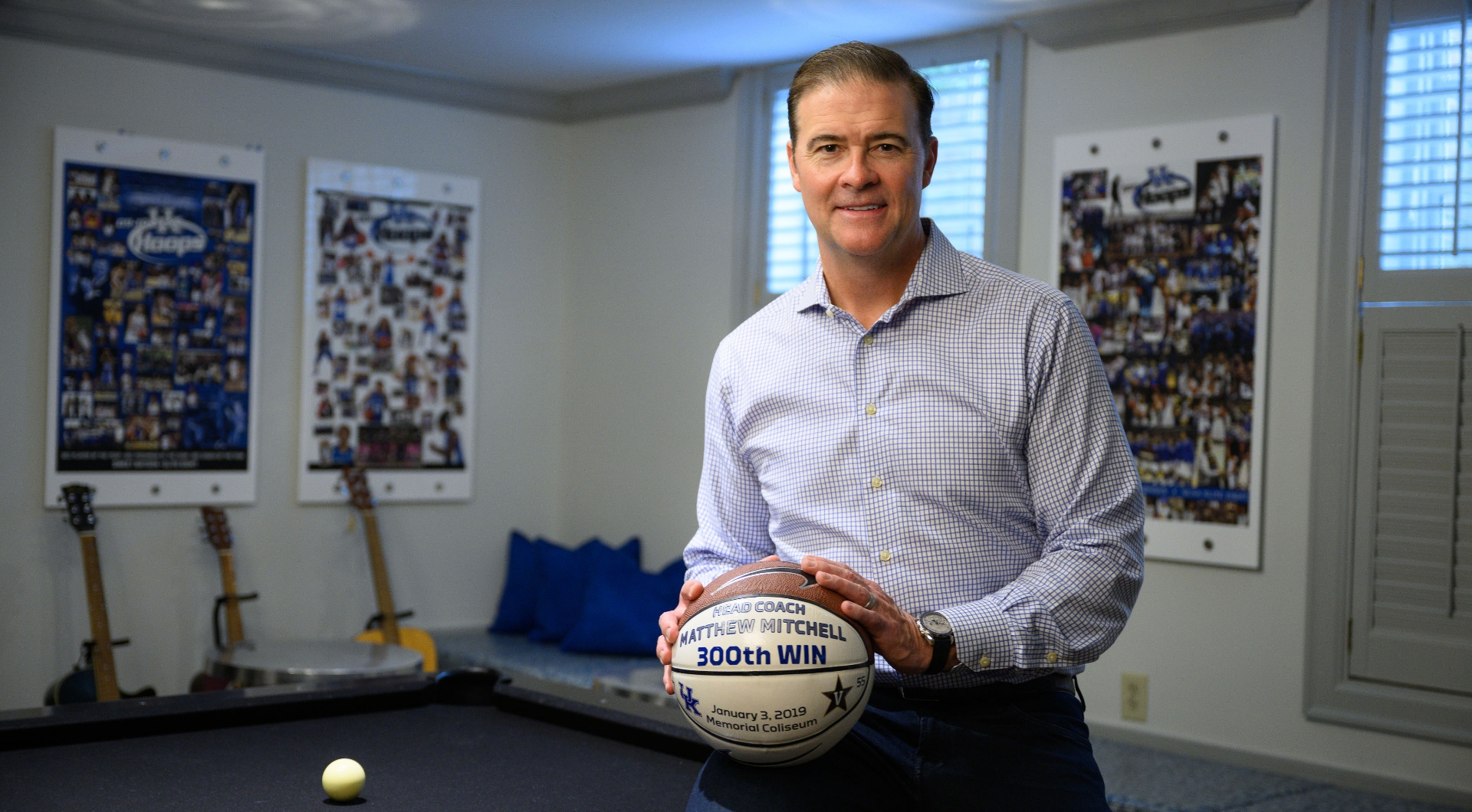 Matthew Mitchell, an older white man with short gray hair, poses in his home with a basketball that celebrates his 300th win. He is wearing a blue and white checkered button-up shirt