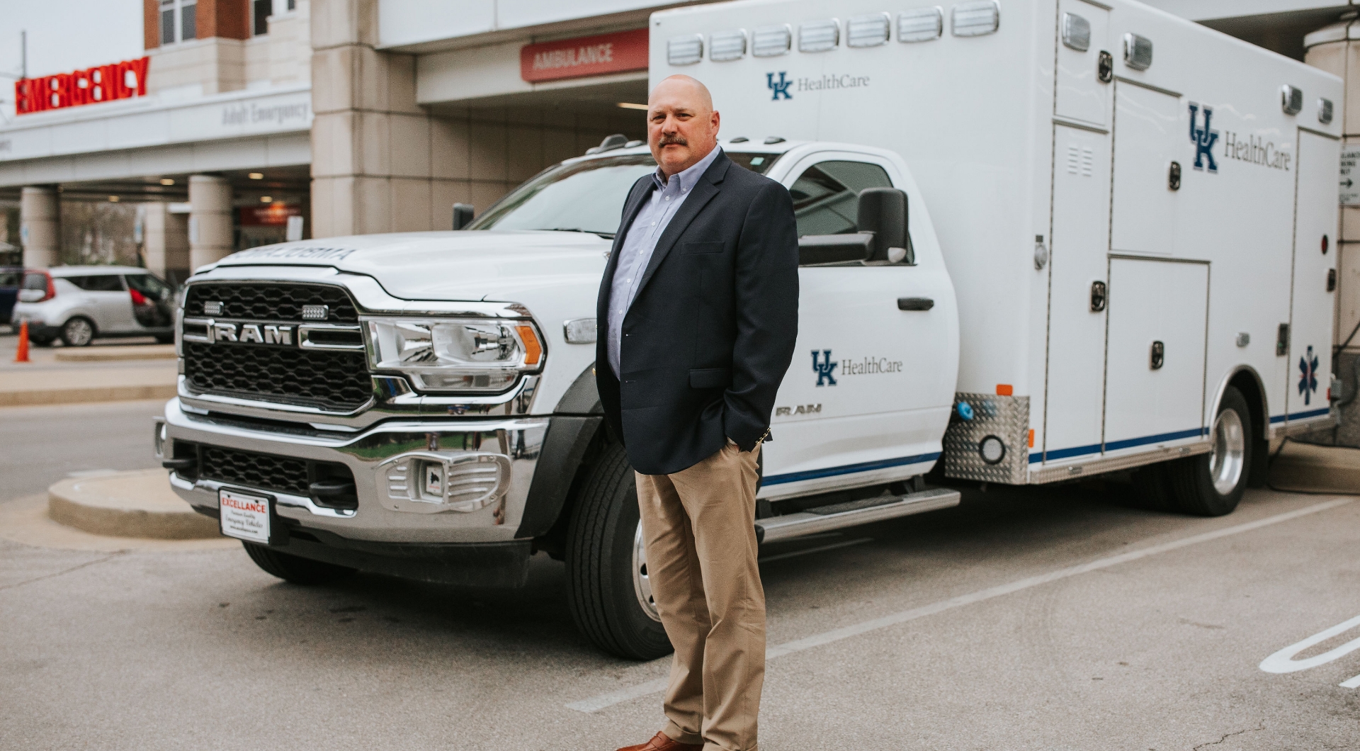 Matt Ward, an older white man with a mustache and shaved head, stands in front of his ambulance with his hands in his pockets. He is wearing a black suit jacket over a gray button-up, and a pair of tan slacks.