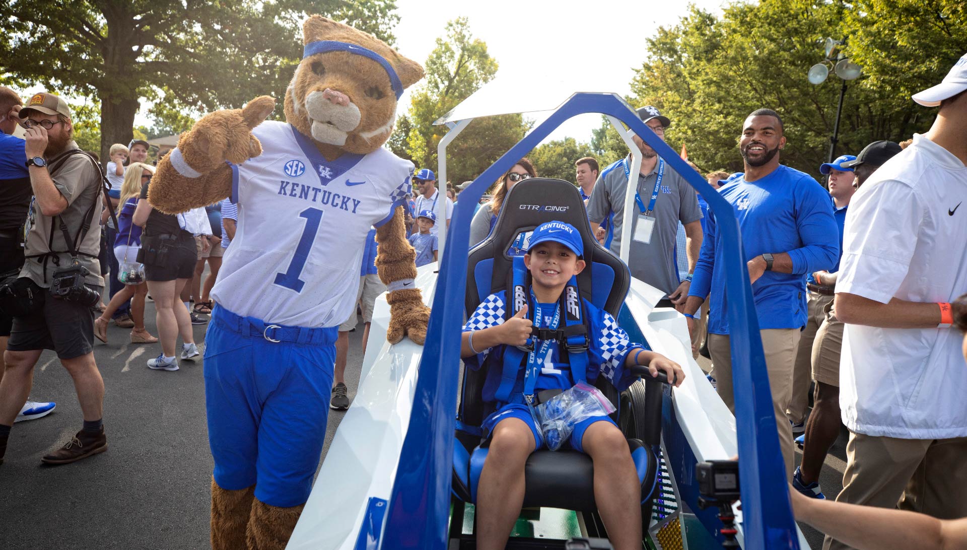 Maximo Shemwell, a young white boy dressed in UK gear, gives a thumbs up while he sits in a futuristic-looking open car. Next to him, the UK wildcat mascot waves.