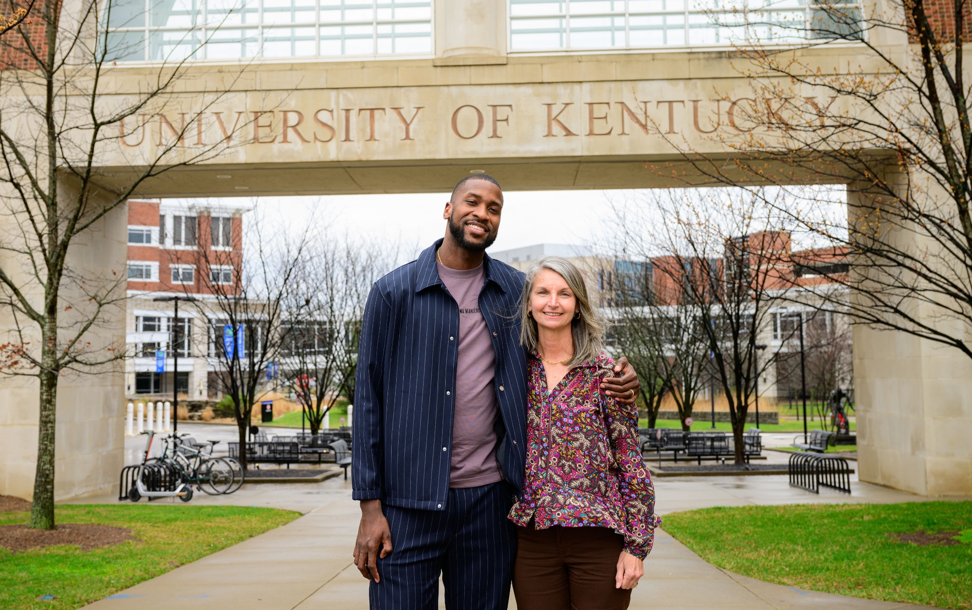 A photo of Michael and Meg Shake smiling for the camera with their arms around each other while on the University of Kentucky campus. Meg is an older white woman with mid-length gray hair. She is wearing a multicolor long-sleeve paisley top and black slacks.