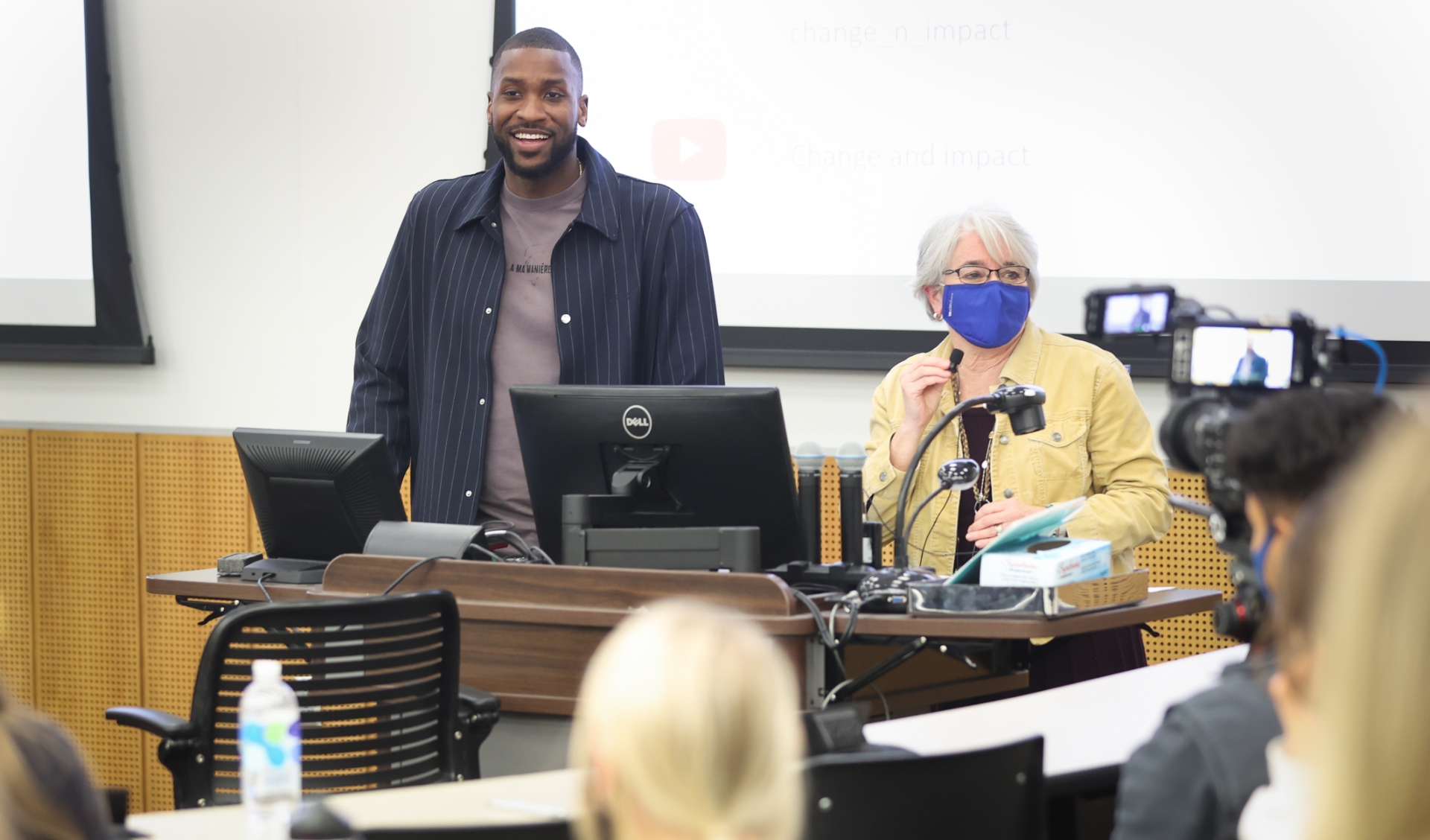 A candid photo of Michael and a UK professor standing at the front of a classroom before giving his presentation. She is an older white woman with short gray hair. She is wearing a yellow denim jacket over a black shirt and gold necklaces.