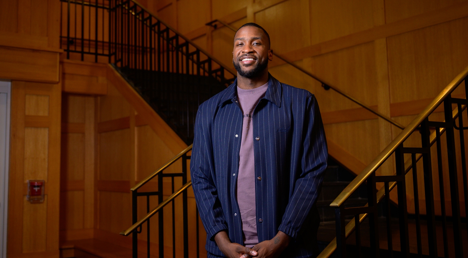 A photo of Michael Kidd-Gilchrist standing in front of a staircase as he smiles for the camera with his hands in front of him. He is a young Black man with short black hair. He is wearing a blue button-down with white stripes over a purple t-shirt.