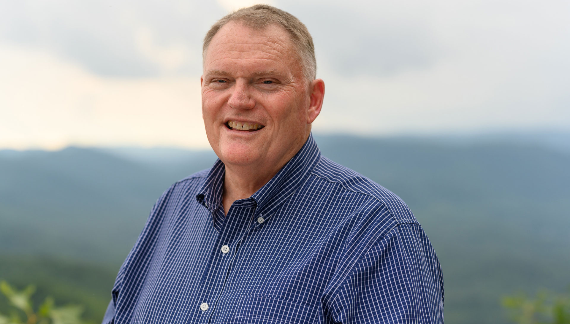 A portrait of Michael Slusher, a middle-aged man with light brown hair wearing a blue and white patterned button up shirt, with mountains behind him.