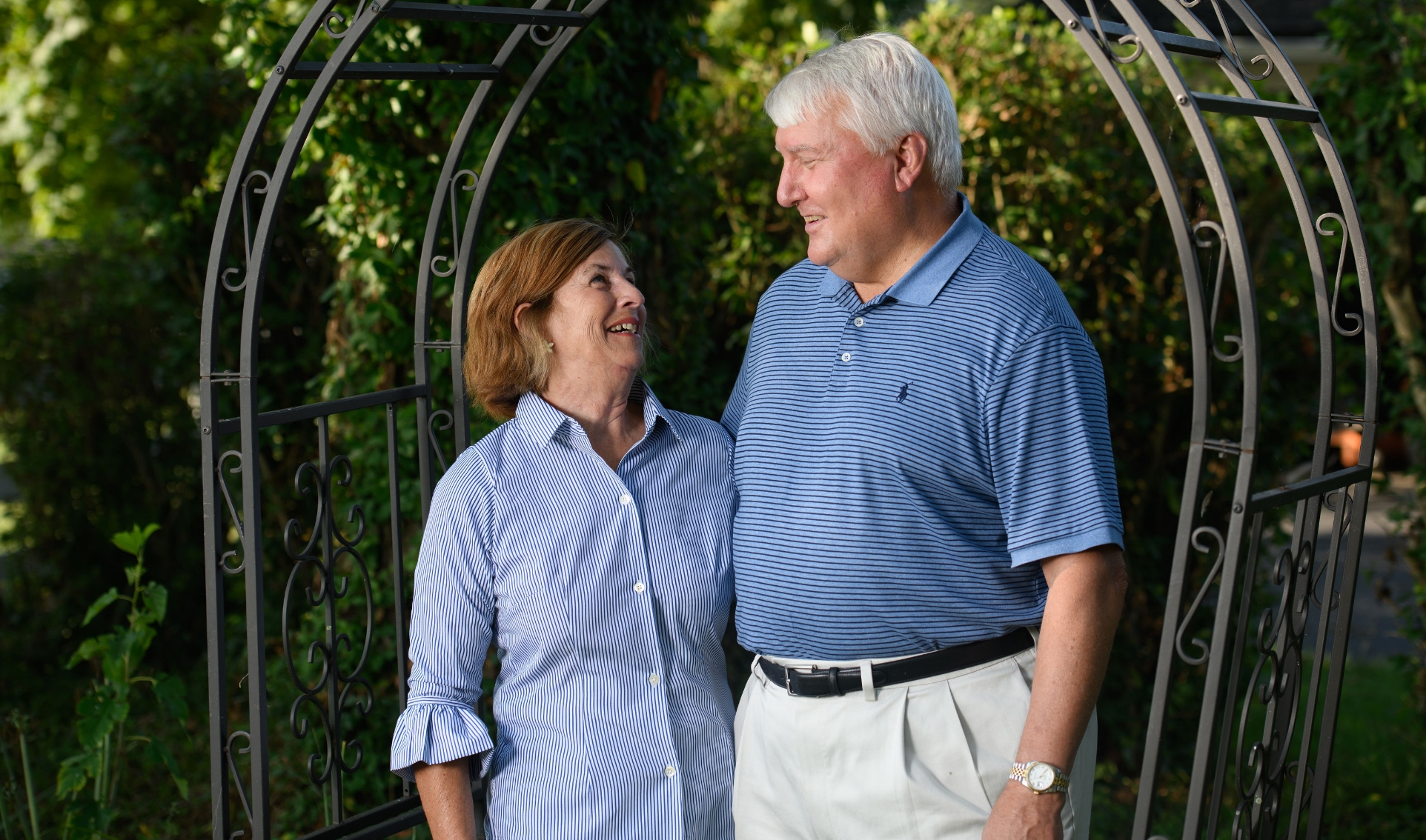 A candid photo of E and her husband smiling at each other while standing under a metal gateway in their garden. Her husband is an older white man with white hair. He is wearing a blue short-sleeve polo that is tucked into his khaki pants.