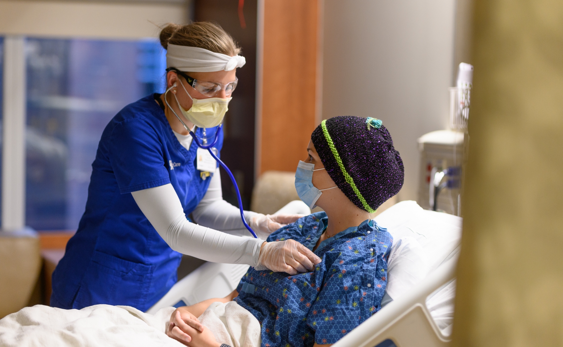 A photo of a nurse holding a stethoscope up to the chest of a patient laying in their hospital bed.