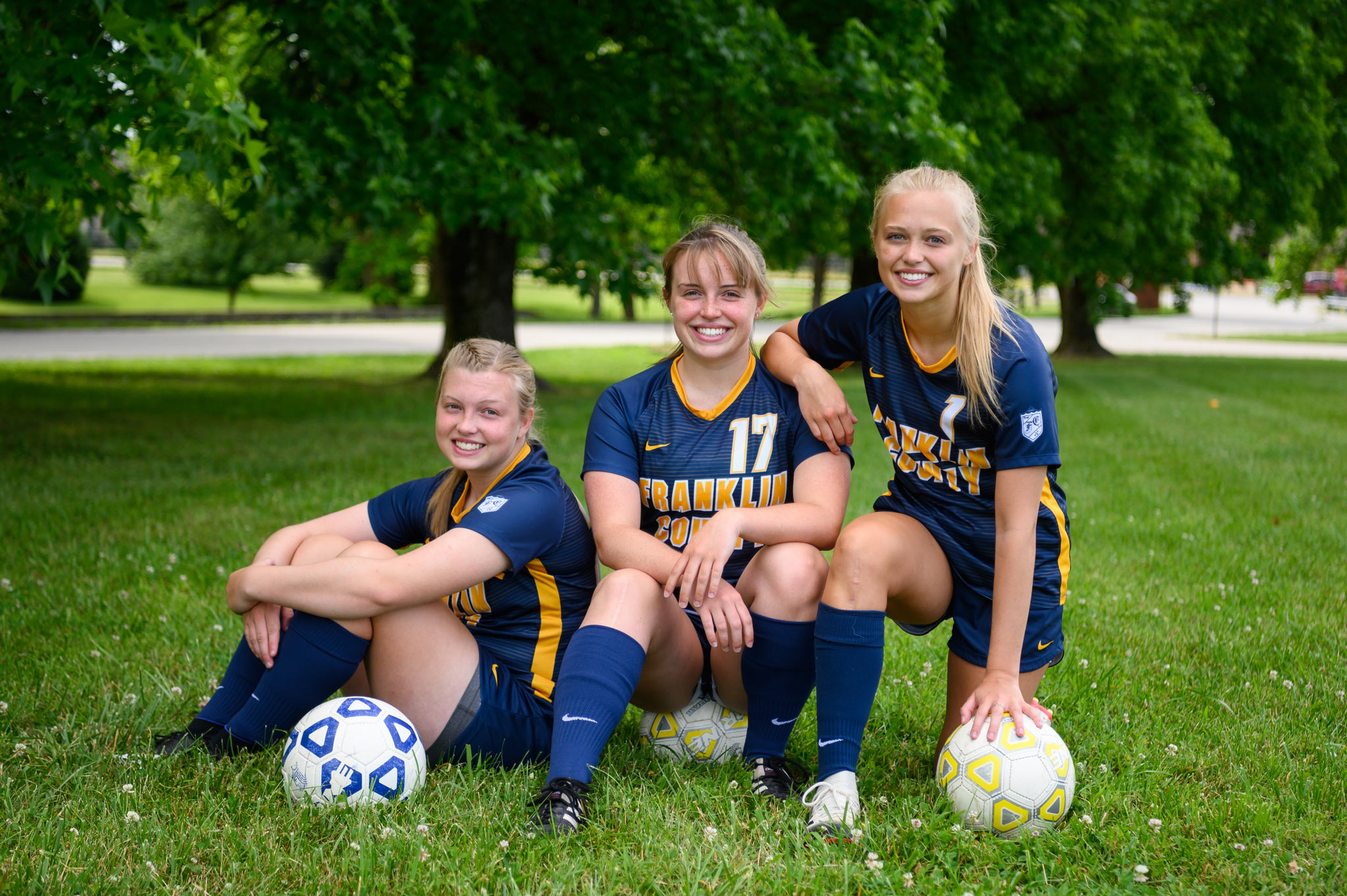 The three girls together crouching down on the field, each with a soccer ball, smiling.