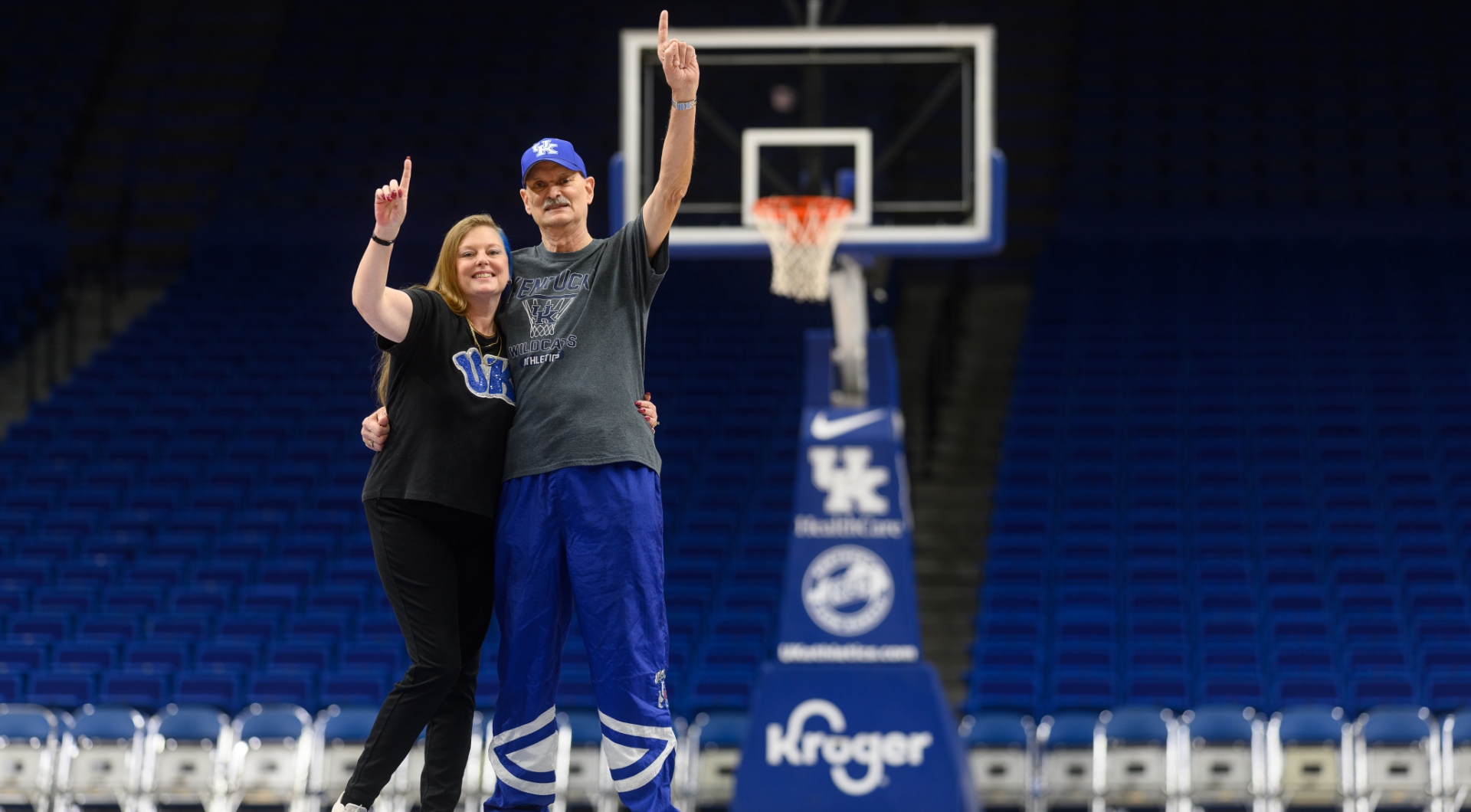 Ronald “Andy” Combs and his wife Julie Combs pose on a UK basketball court with their arms in the air and their index fingers pointed up. Andy is an older white man with buzzed gray hair and is wearing a blue UK hat, a gray short-sleeve Kentucky Wildcats shirt, and blue sweatpants. Julie is a white woman with long, light red hair. She is wearing a dark gray, short-sleeve UK shirt and black leggings.