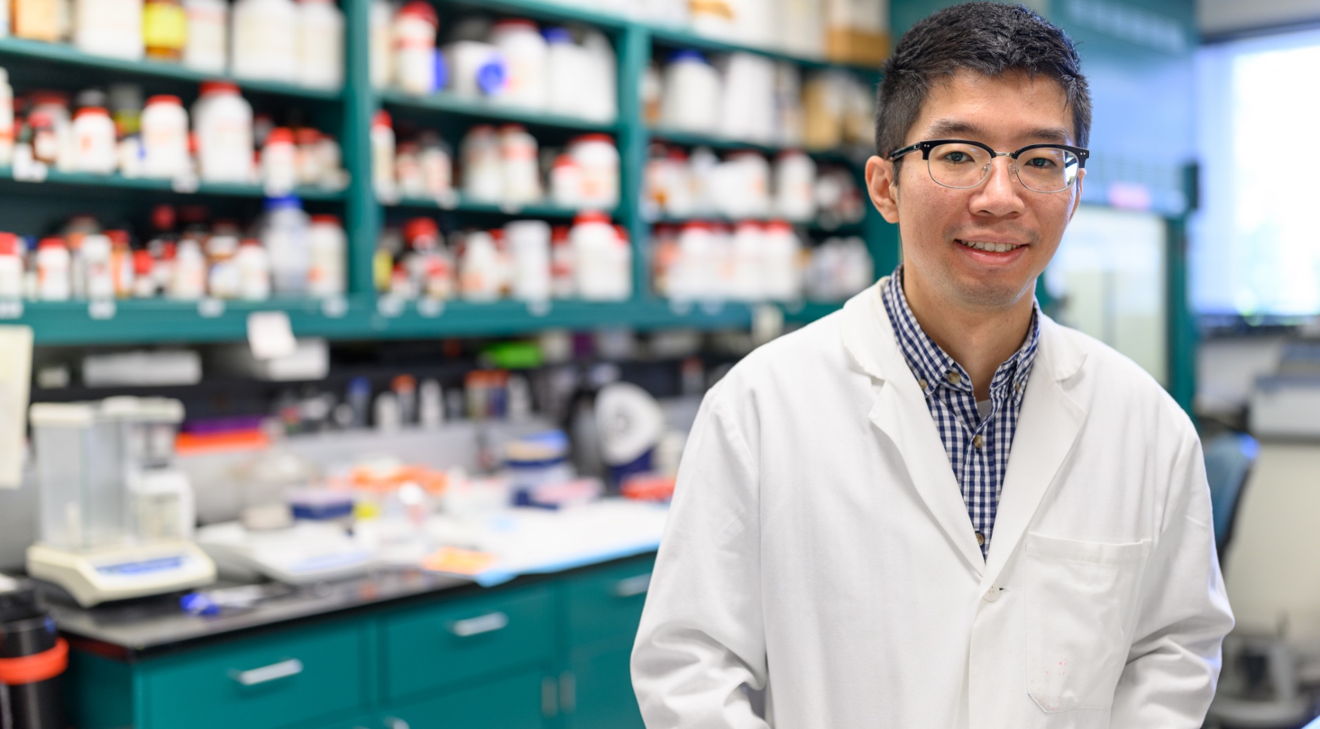 Yueming “Ronnie” Wu, a young East Asian man with short black hair, smiles for the camera as he stands in a laboratory. He is wearing a white lab coat over a blue gingham button-up shirt, and a pair of black-rimmed glasses.