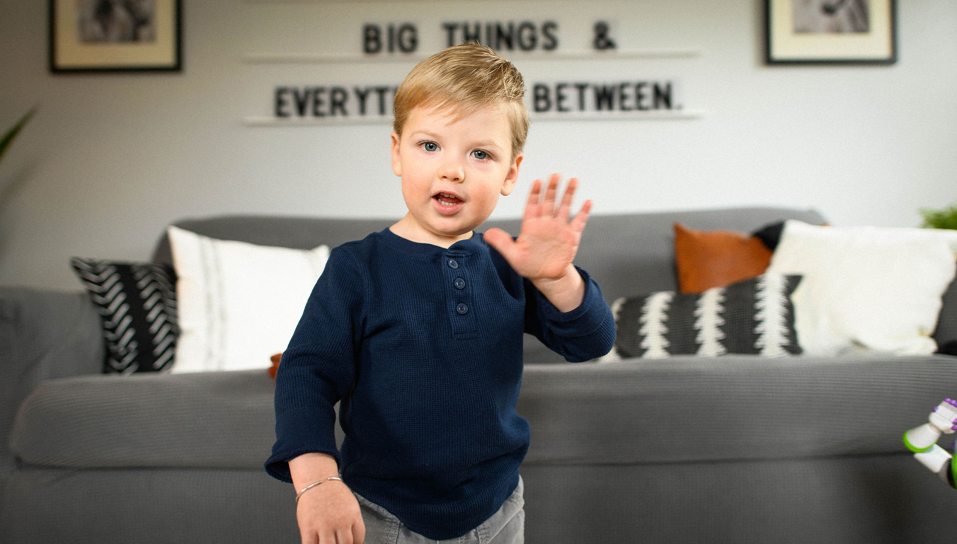 Rory McCamish, a white toddler with blonde hair, blue eyes, and a round face, is standing in his living room waving. He is wearing a navy blue shirt and grey pants.