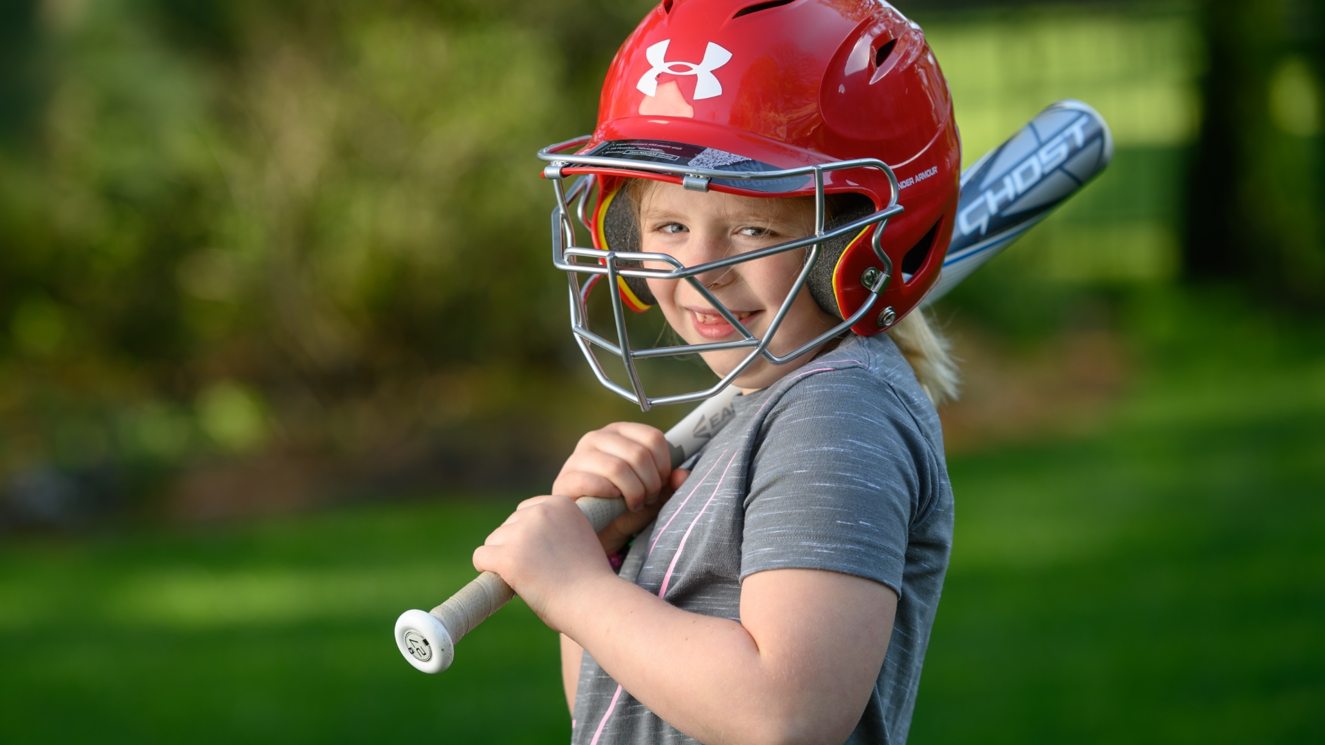 Sarah Beth Ferguson, a blonde eight-year-old white girl wearing a grey shirt, poses outside in a red softball helmet. She holds a grey bat over her shoulder.