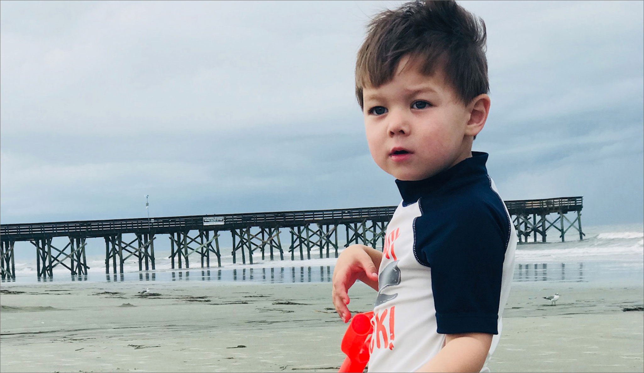 Marco stands on the beach in with a pier in the distance behind him.