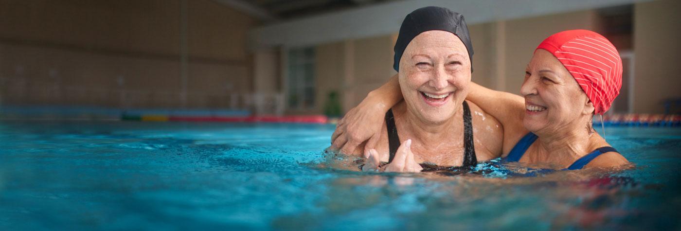 Two older women in swim caps and bathing suits smile and laugh while in a swimming pool.