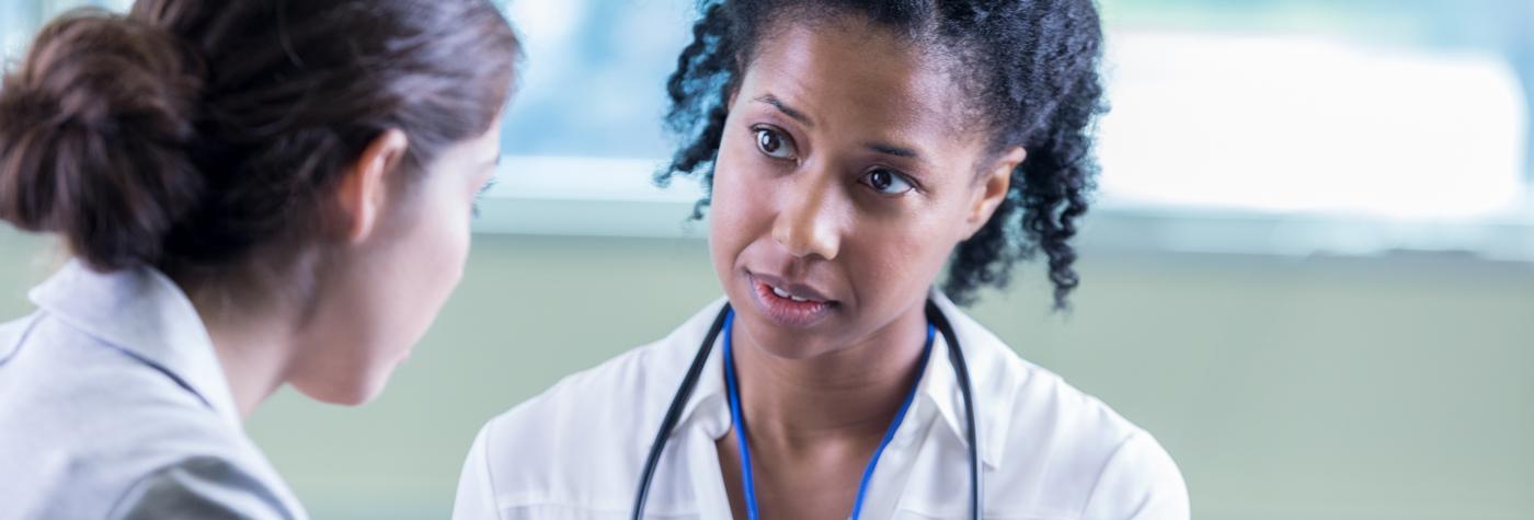 A female provider wearing a white blouse and blue stethoscope around her neck speaks with a patient.