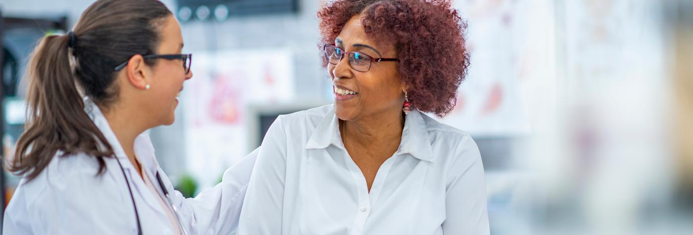 A female provider wearing a white coat and stethoscope and her female patient smile at one another as they have a conversation. 