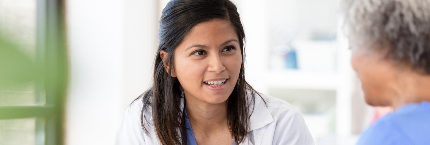 A woman in light blue scrubs and a white coat speaks to a female patient.