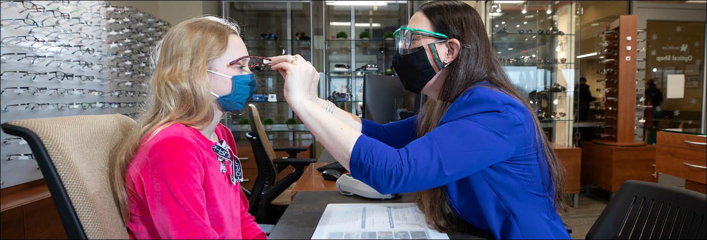 A member of the Optical Shop team adjusts a pair of glasses on a customer. The two people pictured, both women, are wearing face masks.