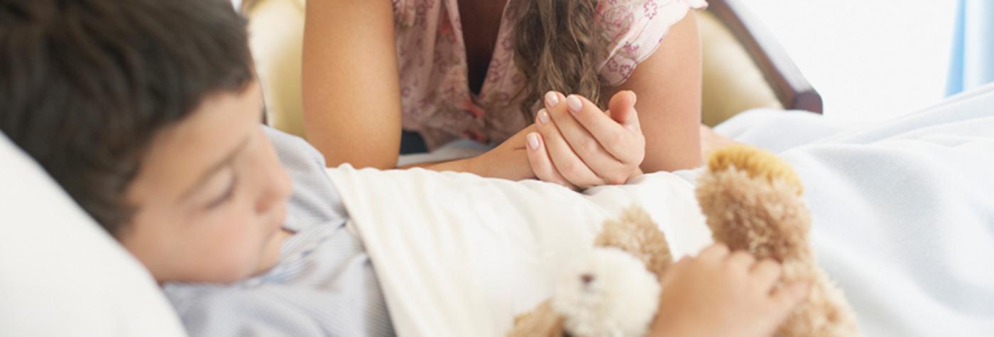 Young boy sleeping in hospital bed with teddy bear and mother looking over him