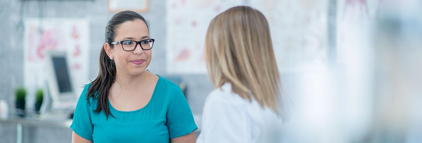 A woman in a teal blouse talks to her doctor, also a woman, who is shown from behind.
