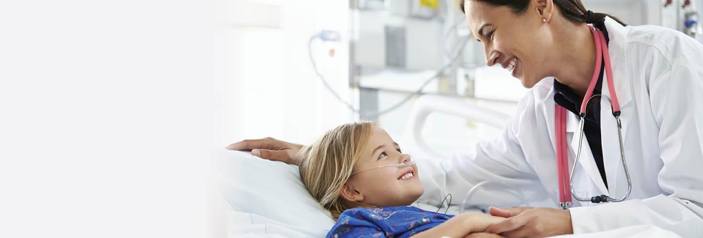 A female doctor in a white coat smiles and talks with a blonde-haired child, dressed in a blue hospital gown, who is resting  in a hospital bed.