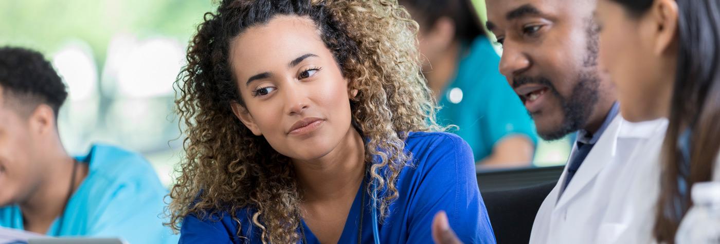 A young woman in blue scrubs listens to a male preceptor, dressed in a white coat, explain a concept in a classroom setting.