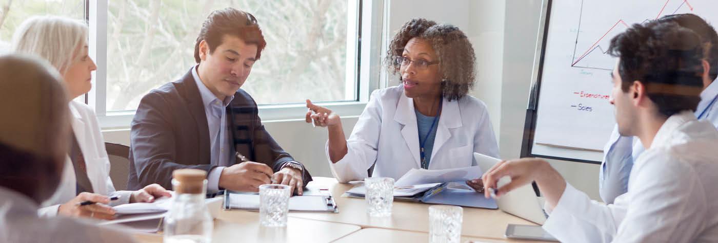 A group of providers wearing white coats and people in business attire gather around a conference table. 