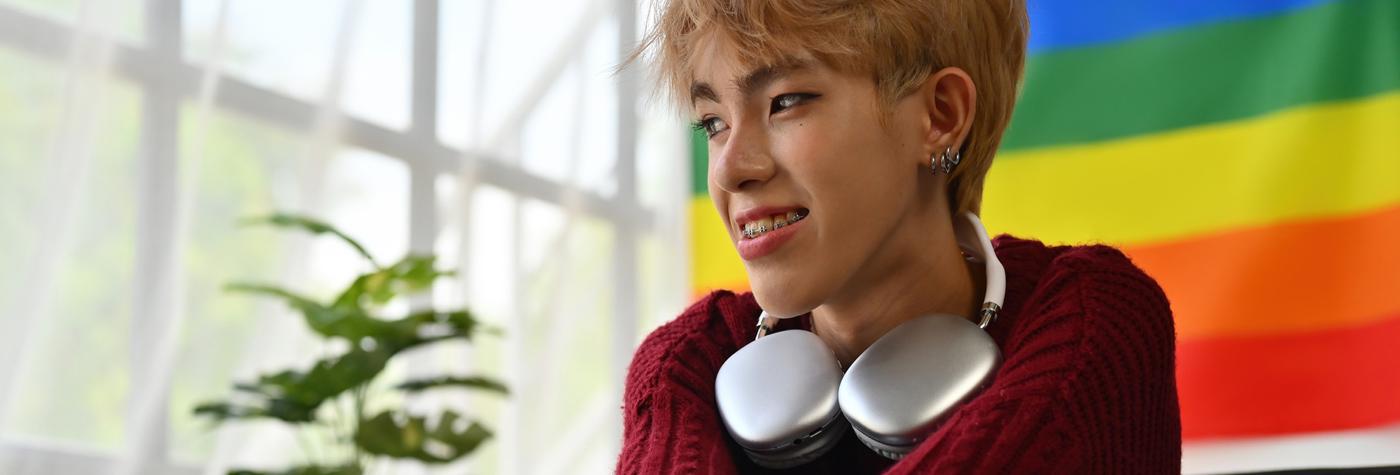 A young adult wearing a dark red sweater with silver headphones around their neck sits in front of a Pride flag.