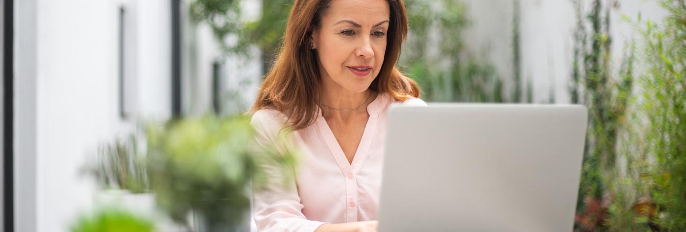 A woman in a light pink blouse types at a laptop. She is surrounded by potted plants.