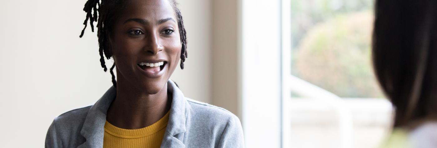 A Black woman wearing a yellow tee shirt paired with a grey buttondown smiles as she speaks with another woman, whom we only see from behind. They are both seated next to a window.