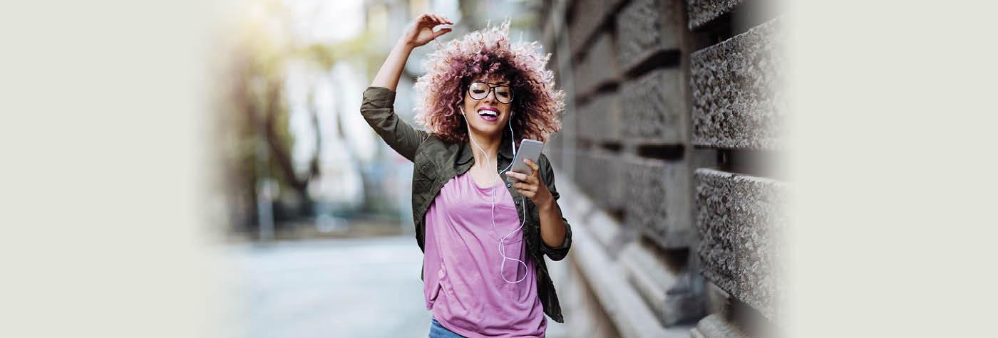 A young woman wearing a pink tee shirt and green jacket walks outside while listening to music and joyfully pumping her right fist into the air.