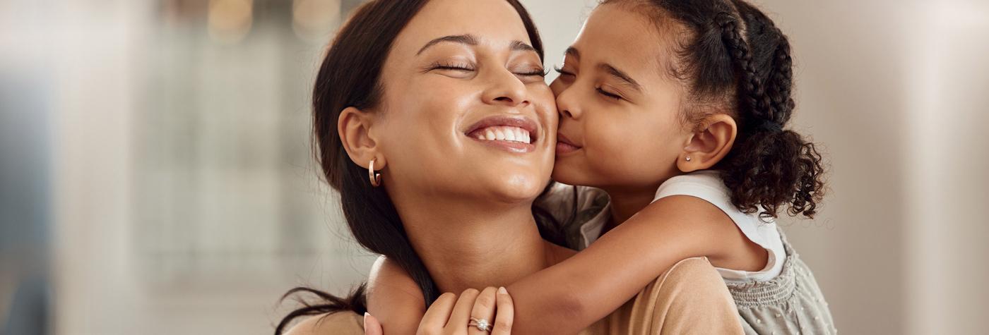 A young mother smiles as her preschool age daughter embraces her from behind and kisses her cheek.