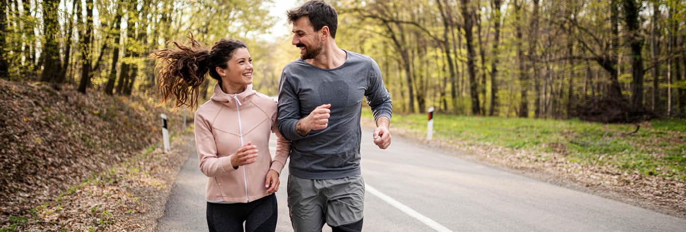 A smiling young couple jogs together on a road with woods on either side of them.