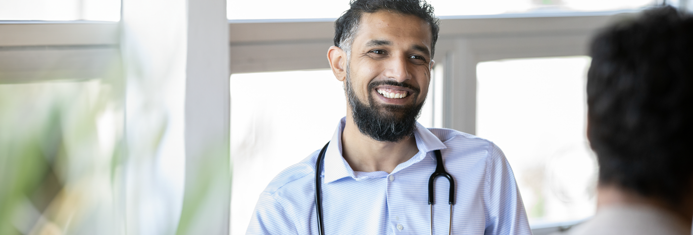 A man wearing a light blue polo shirt with a stethoscope smiles as he speaks with a patient.