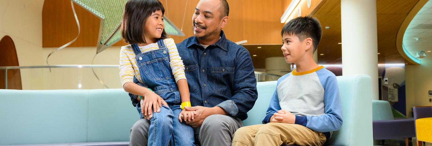 Father with son and daughter sitting on a light blue couch in a hospital lobby