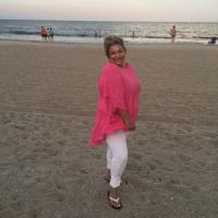 Katrina Thompson wears a bright pink shirt, white slacks and sandals as she stands on the sand with the ocean behind her.