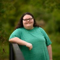 A photo of Abby smiling directly into the camera as she poses with one elbow leaning against a metal railing.