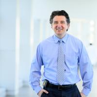 A photo of Dr. Samir Softic smiling for the camera with his hands in his pockets as he stands in a hospital hallway. Dr. Softic is a Bosnian man with wavy salt-and-pepper hair. He is wearing a blue long-sleeve button-up shirt with a light blue patterned tie.