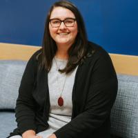 A photo of Terra Lucas sitting on a gray couch as she smiles for the camera. Terra is a young white woman with long straight dark-brown hair. She is wearing a pair of dark-rimmed glasses, a black cardigan over a gray shirt, and a gold necklace that has a large reddish brown gem.