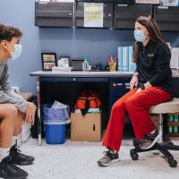 AJ sits on the bench across from his school nurse. His nurse, middle-aged white woman with long, straight brown hair, sits on a chair, wearing a black sweatshirt and red-orange pants. Both she and AJ are wearing masks.