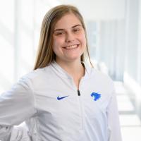 Allsion stands in a white hallway with a wide smile, wearing a white jacket with the University of Kentucky athletics emblem on the chest.