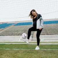 Arin Wright balances a soccer ball on one foot while standing in the goal net. She is visibly pregnant and happy.