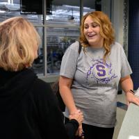 Callie stands at the front desk of the aquatic center and smiles as a blonde woman with short hair and a dark hoodie holds her wrist.