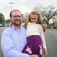 A photo of Cate smiling as her father Jerry holds her in his arms. Jerry is a white man with short blond hair, and he is wearing a pair of black-rimmed glasses and a blue checkered button-up shirt.