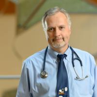 Dr. Stefan Kiessling, an older white man with short gray hair, looks directly into the camera with a warm expression on his face. He is wearing a long-sleeve blue button-up with a blue tie, and a stethoscope around his neck.