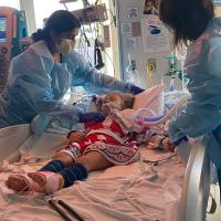 A picture of Cate in her hospital bed. She is hooked up to several different machines, and has two attending nurses standing over her.