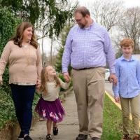 A candid photo of the Cook family holding hands and smiling as they walk down the sidewalk together. Cate’s mother Ann, a white woman with long brown hair, is on the far left. She is wearing a light brown sweater over a white button-up, and a pair of blue jeans. Cate’s brother Charlie (far right) is a young white boy with medium-length red hair. He is wearing a blue long-sleeve button-up and a pair of khakis.