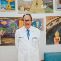 Dr. Carl Backer, an older man with thin glasses wearing a white doctor’s coat, smiles in front of a wall of children’s paintings.