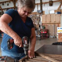 A close-up photo of Charlotte, wearing a navy blue shirt and jeans, using a power drill on a piece of wood in the barn on her farm.